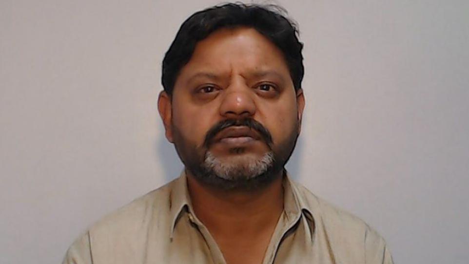 A mugshot of Kasir Bashir shows him staring directly at the camera with no expression on his face. He has black hair, a black mustache and a greying beard, and is wearing a beige shirt.