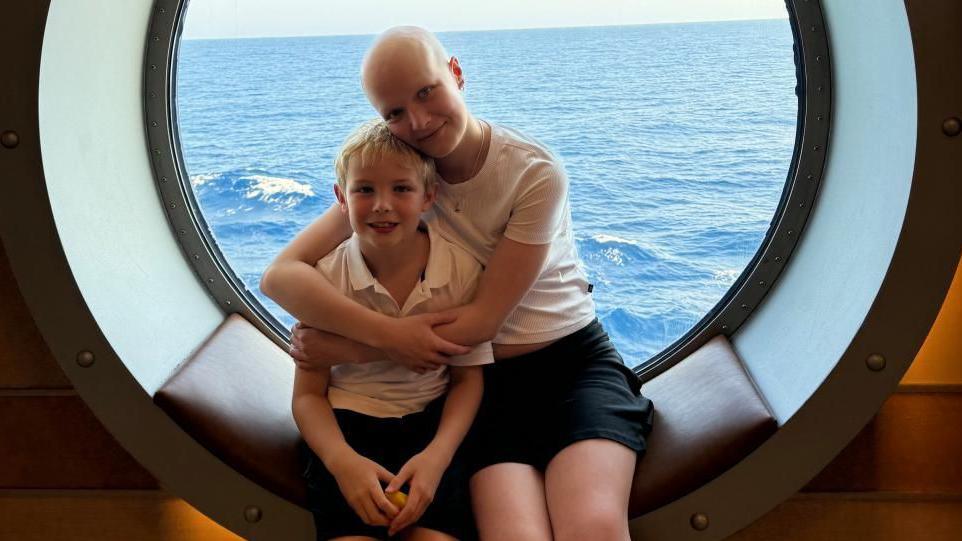 Mateo and Liz sit on a padded seat in the window of a ship. Liz has her arms around Mateo and they are both smiling at the camera.