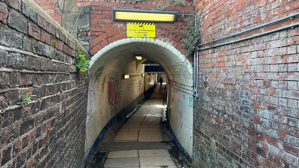 A rundown subway built out of red brick, with its white brick internal walls covered in graffiti. It has a tiled floor, and someone can be seen walking at the far end of the tunnel.