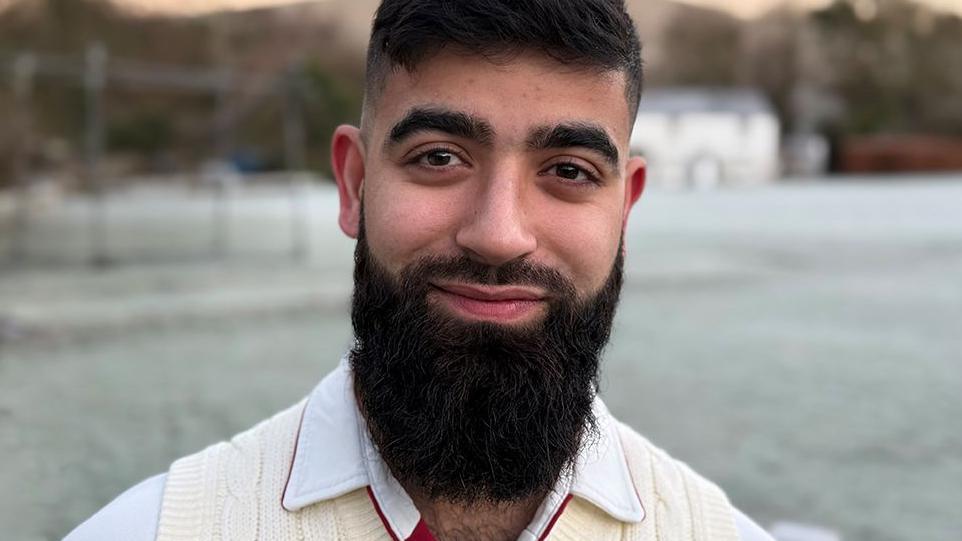 Head and shoulders image of Usmaan Akram standing on a frost covered cricket pitch. He is smiling, has dark hair and a long dark beard and is wearing cricket whites