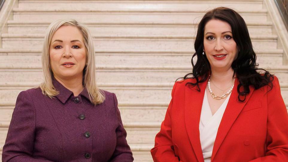 Michelle O'Neill and Emma-Little Pengelly stand side by side with the sandy stairs in Stormont behind them. O'Neill is wearing a dark purple button-up blazer, with shoulder length blonde hair and has makeup on. Emma-Little Pengelly is wearing a red blazer and a white top with a pearl and gold design necklace. She has long black hair and red lipstick on.