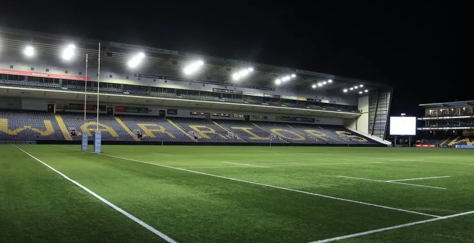 A rugby stadium at night viewed from the pitch, lit up by floodlights. Seats in the stand are coloured to spell out "WARRIORS".