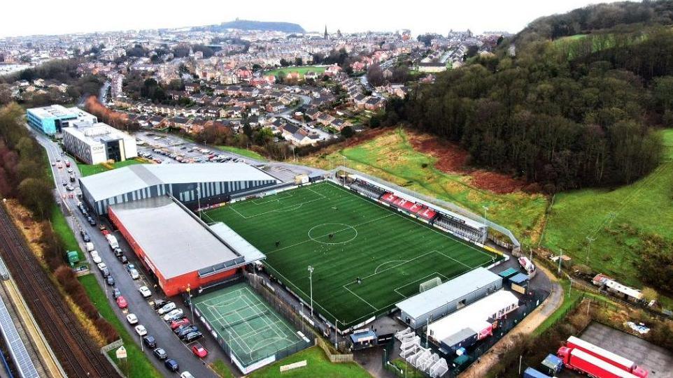An aerial view of the football pitch, with an array of buildings in the background and cars parked next to the stadium.