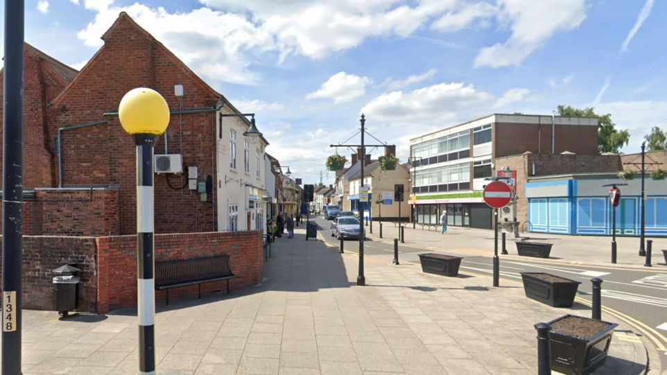 A view of a street with pelican crossing lights, a row of shops and street lighting with pavements on either side