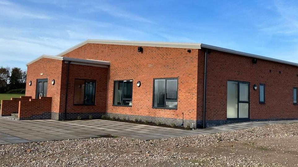 A large new built red brick building, single storey with black framed windows and a porch extension on the front