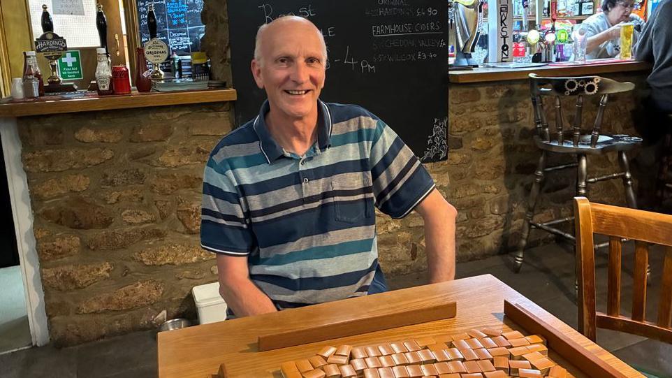 Mark Trenchard set up Shipham's Mahjong club. He is a slim, middle-aged white man who is wearing a blue striped top. He is sitting in a pub with a Mahjong table in front of him.