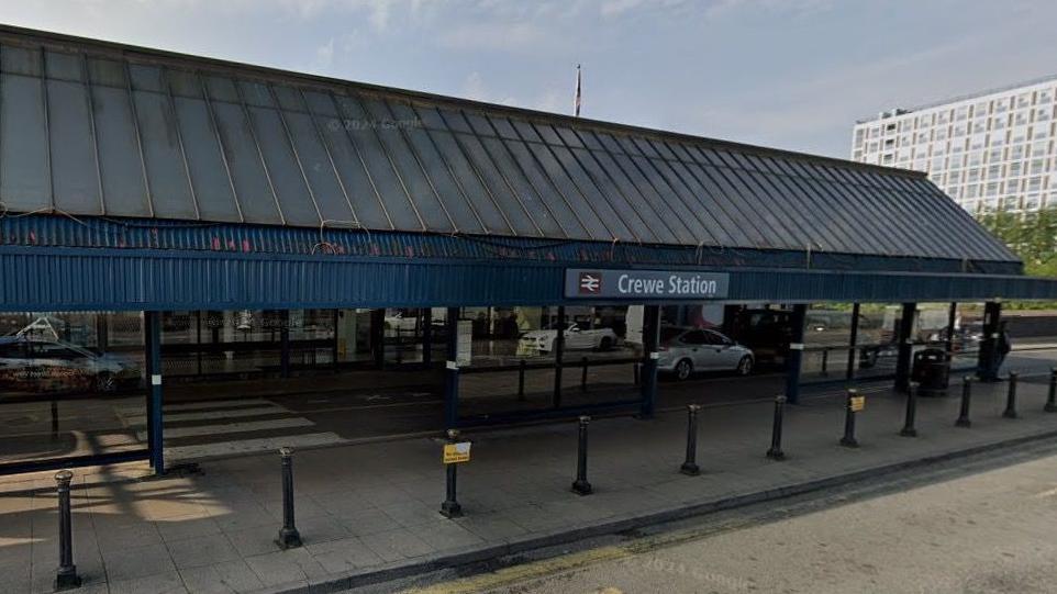 The outside of a railway station. It is a single storey building with a blue slanted roof and a sign that says Crewe Station. There are bollards lining the pavement in front of the building