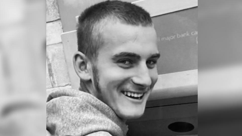 A black and white image of Ben Moncrieff. He is wearing a hoodie and looking over his right shoulder towards the camera with a wide smile. 
