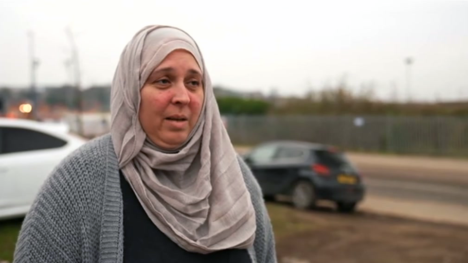 A woman standing outside, with a road and cars in the background. She is wearing a pale headscarf, black top and grey cardigan.