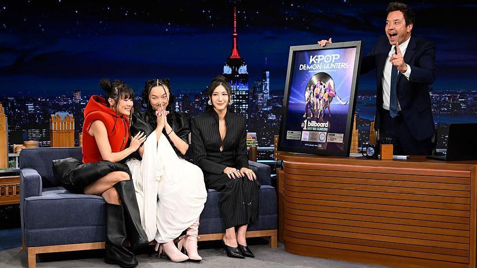 Rei Ami, Audrey Nuna, and EJAY, who provide the singing voices of Huntr/x. They performed on Jimmy Fallon's US talk show. Fallon is holiding a platinum disc with which the singers are being presented.