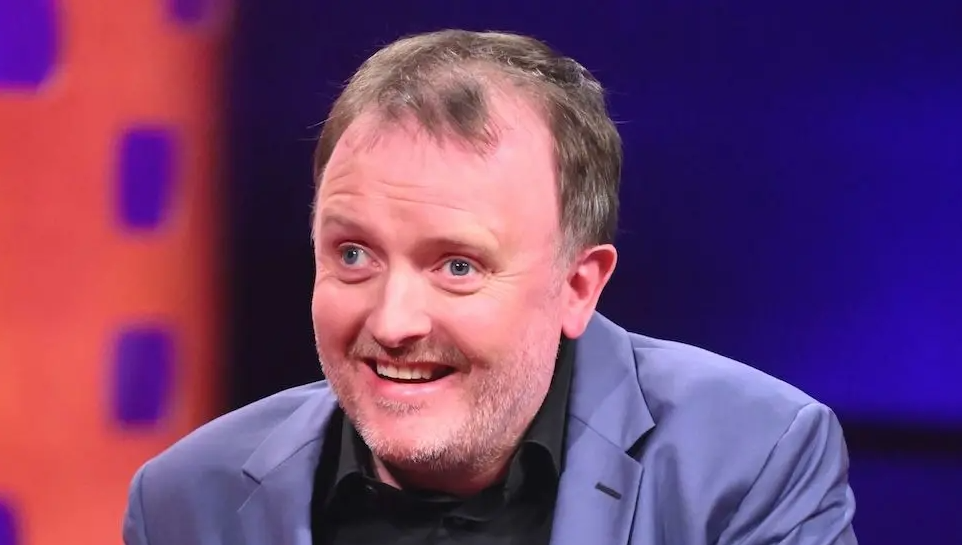 Comedian Chris McCausland, wearing a black shirt and powder blue jacket, smiles at someone or something off-camera.