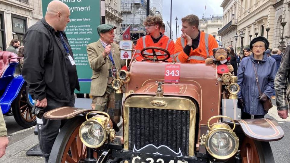 Two young men in orange hi vis jackets sit on a vintage car on Pall Mall as they listen to a man with a flat cap and a tweed blazer on.