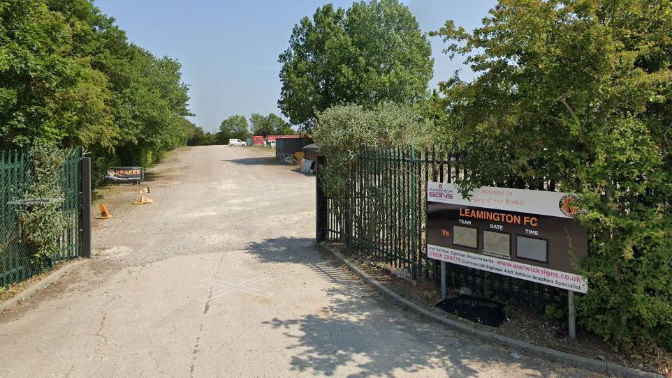 The entrance to Leamington FC's current ground. Metal fencing is on each side as well as trees and sign with the words Leamington FC written at the top in orange.
