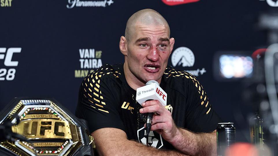 Sean Strickland, wearing a black t-shirt with gold pattern on the shoulders, sits talking into a white and black microphone, held in his left hand, with a gold octagonal UFC belt to his right