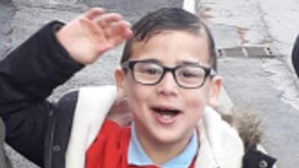 little boy with gelled down brown hair and black square framed glasses. He is smiling and waving 