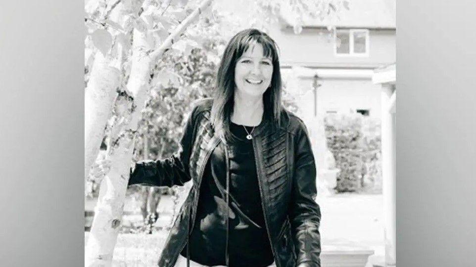 A black and white image of a smiling woman with shoulder-length hair wearing a leather jacket and necklace.