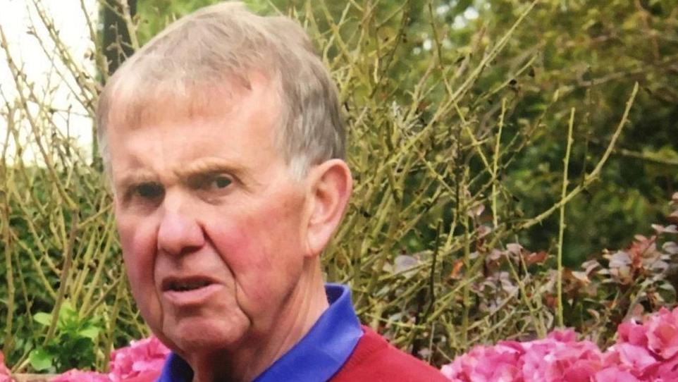 Malcolm Flynn. He is an older man with light hair. He is wearing a blue-collared shirt under a red jumper. He is standing in a garden surrounded by pink flowers and greenery.