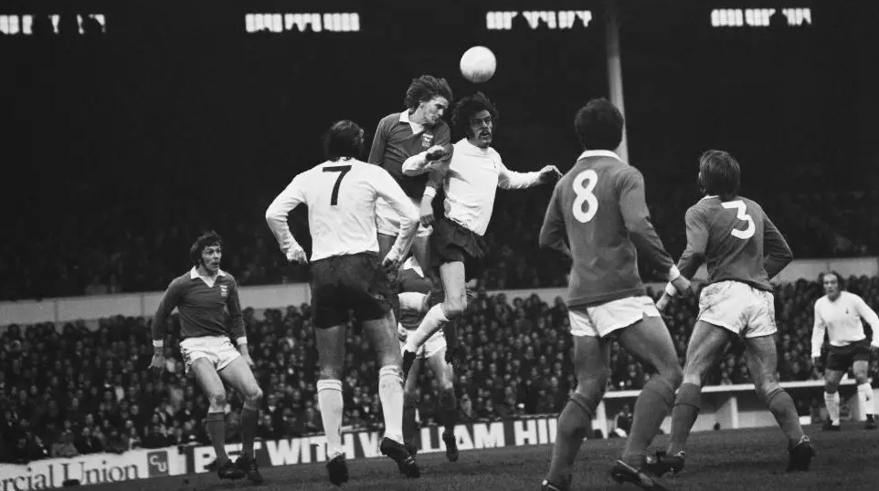 A black and white image showing two footballers contesting a header on the pitch as other players look on.