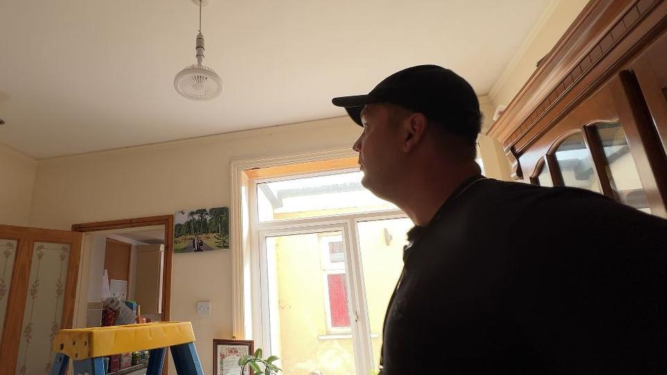 Mr Waines is in a living room, he is wearing a black shirt and a black cap, he is looking at a fan light which is attached to a bulb fitting and is hanging from the ceiling. He has his step ladder to the left and is preparing to remove it.