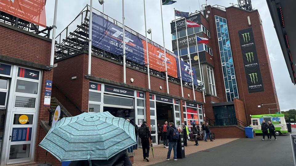 People standing at the back of the TT grandstand, which is a redbrick building with a rectangular tower. One of the spectators has a light blue umbrella up.