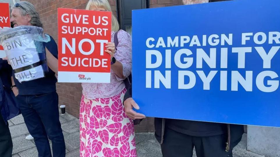 People holding up signs for and against the bill. They include a blue sign with the words Campaign for Dignity in Dying, and a red sign with Support Not Suicide written on it. There is also a waste paper bin with the words Bin the Bill on it.