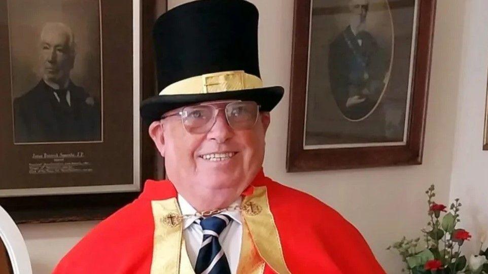 A man wearing a black top hat with a yellow buckled ribbon across the middle and is wearing a red cloak over a white shirt and blue striped tie with a golden clasp fashioning the cloak.