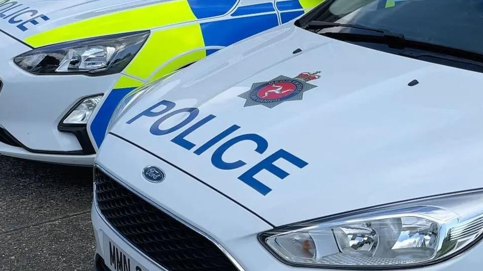 The bonnets of two police cars parked next to each other. The nearest has the word police in blue writing on a white background, and features the Isle of Man Constabulary's emblem. Blue and yellow paint can be seen on the side of the other car.