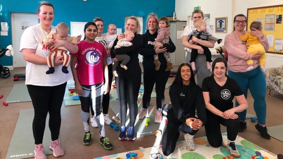 A group of seven women in an exercise class with their babies. There are two instructors. They are all wearing work out gear and are smiling at the camera. 