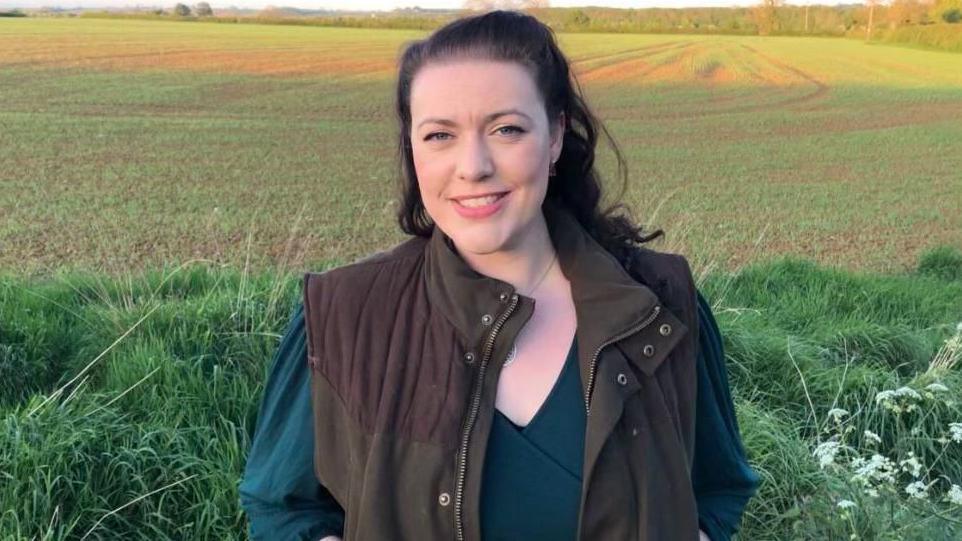 Alicia Kearns MP is wearing a brown body warmer and dark blue long-sleeved top. She has long brown hair slicked back and is smiling into the camera. Behind her is a grass verge and then a farmed field behind that.