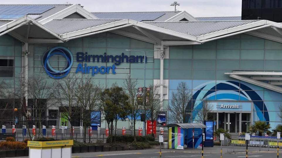 An exterior of Birmingham Airport, with the name and logo in blue on the front of a glass building which also says arrivals. Small trees and a road are visible in front of the building, as well as a bus stop.