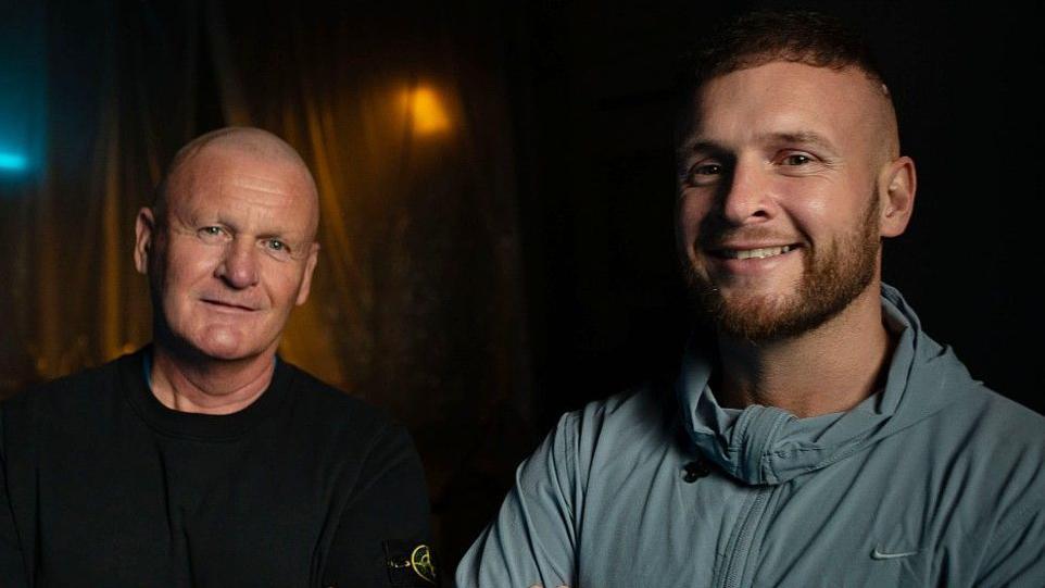 Andrew and Macaulay Dodd pictured standing next to each other. Both looks at the camera and smiles. Andrew wears a black top and Macaulay a light blue sports zip-up top. It is a head and shoulders shot of them.