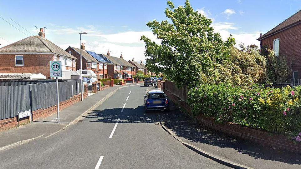The start of Stainforth Avenue, Bispham, Blackpool. A mini is parked on the right of the street in the shadow of trees from a garden. It is a sunny day. There is a 20 mph road sign saying 'Twenty's Plenty'.