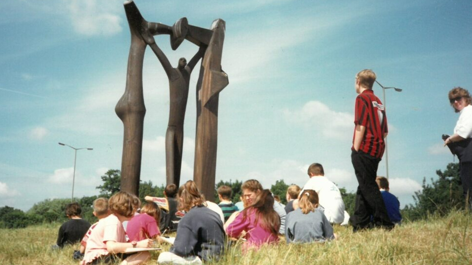 A group of children sat on the grass while on a school trip to see the Peterborough Arch sculpture in 1998. The sky is blue with some clouds and the grass is long and looks quite dry. The sculpture stands tall in front of the children and shows an abstract wooden man with his arms up, touching the top of a wooden arch.