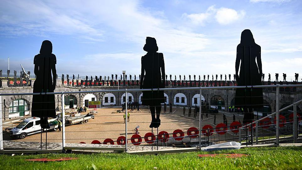 3 silhouetted figures of women in the foreground, with the stone walls of Nothe Fort in the background lined with similar figures of armed forces personnel around its circumference, with the gates lining the walls decorated with red poppy wreaths.