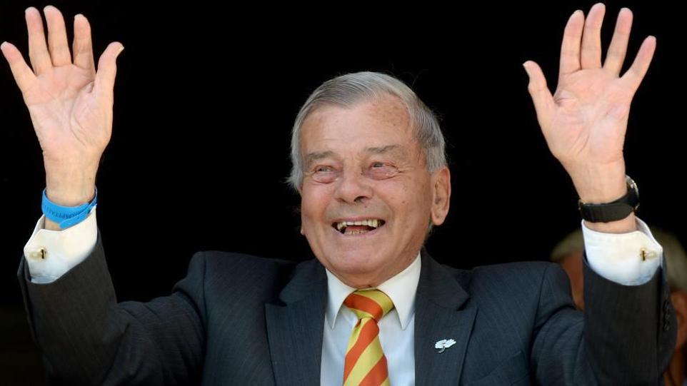Dickie Bird, an elderly man with short grey hair, dressed in a dark suit, white shirt, and a striped yellow and red tie. He is smiling broadly and raising both hands in the air. The background is dark, with other people partially visible behind.