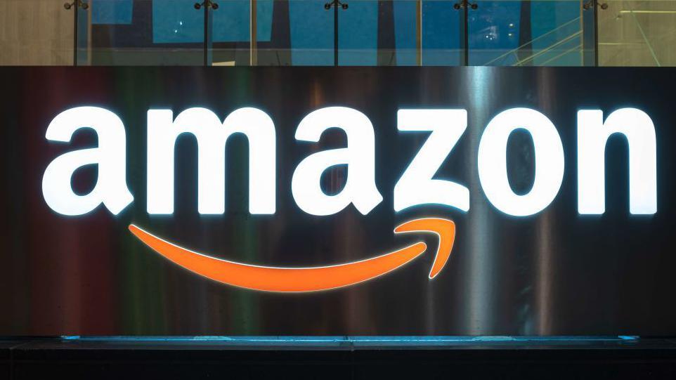 Illuminated Amazon sign on the side of a building with the company's orange arrow under the company name.