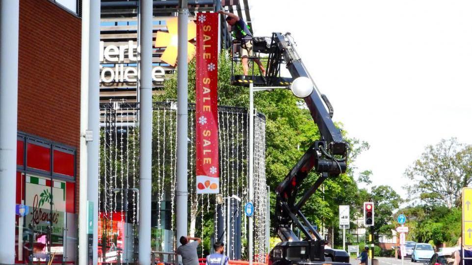 A man on a cherry picker is fixing a sign advertising a sale to the outside of West Herts College near some Christmas lights