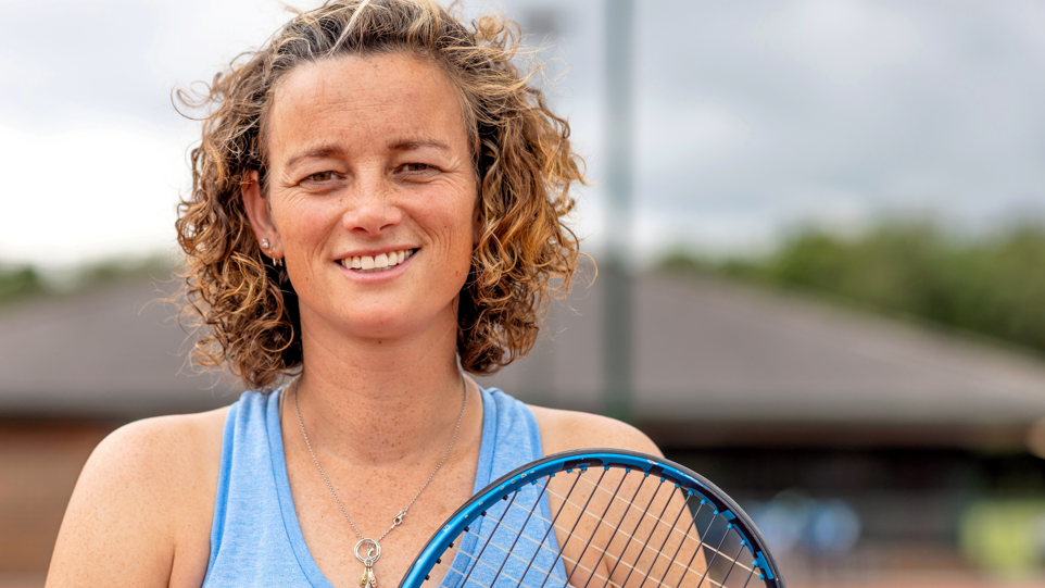 A professional close-up shot of Catherine, smiling to camera, wearing a blue sports vest and holding her tennis racket up near her shoulder. She has very curly brown hair. The background of the tennis court is blurred.