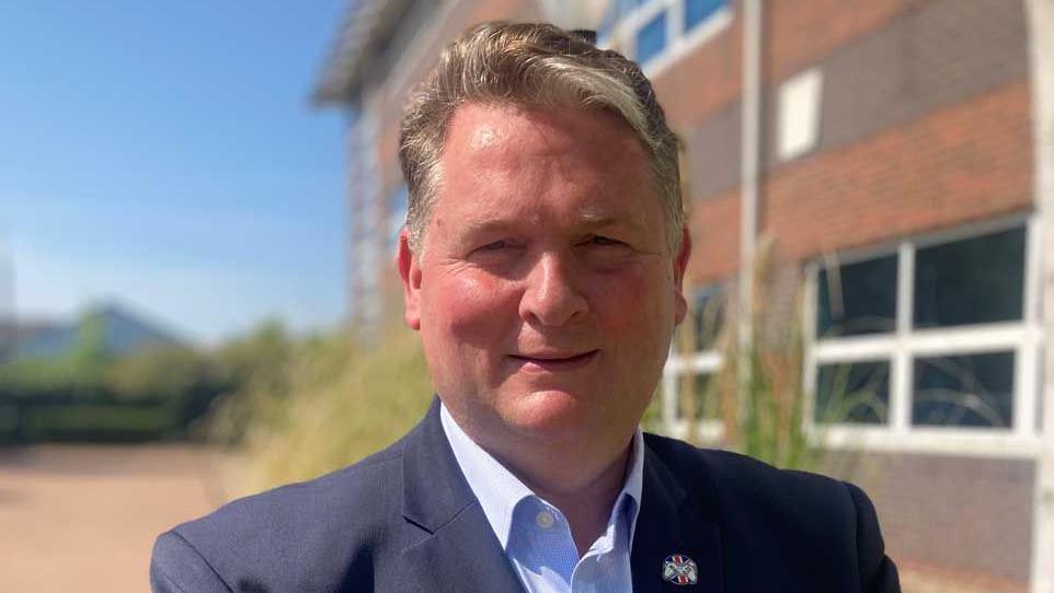 Image shows man wearing blue suit looking into the camera on a sunny day outside a building.