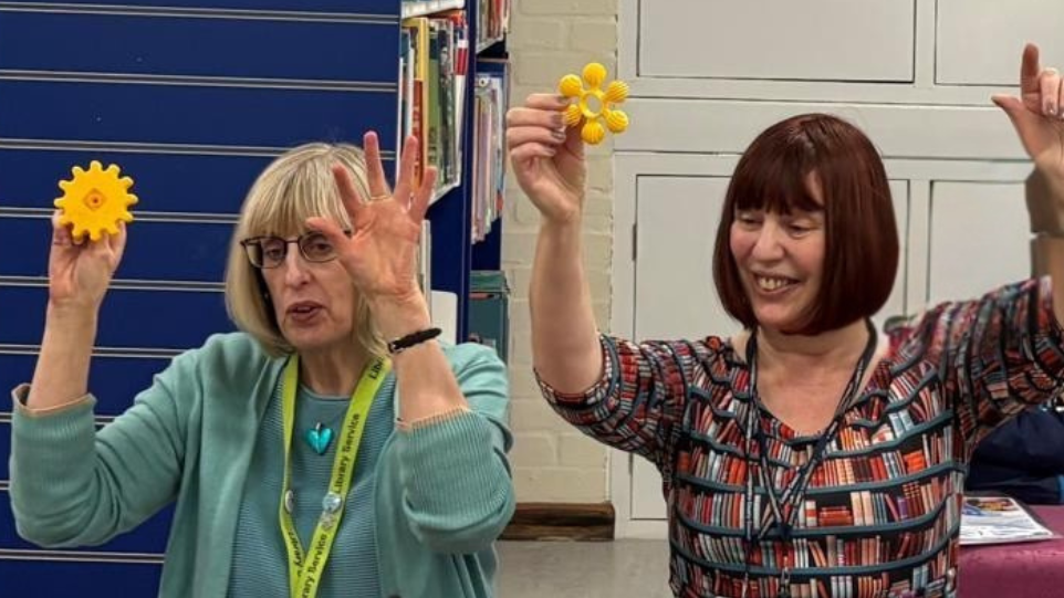 Two women sat on a chairs in a library with their arms in the air while holding what looks similar to a yellow flower in their left hand. They are telling a story to some children (not pictured). The woman on the left has blonde hair and a fringe styled into bob. She wears a turquoise top, matching love heart necklace and glasses. The woman on the right has red/brown hair styled into a bob with a fringe. She is wearing a dress that has rows of bookshelves printed onto it. 