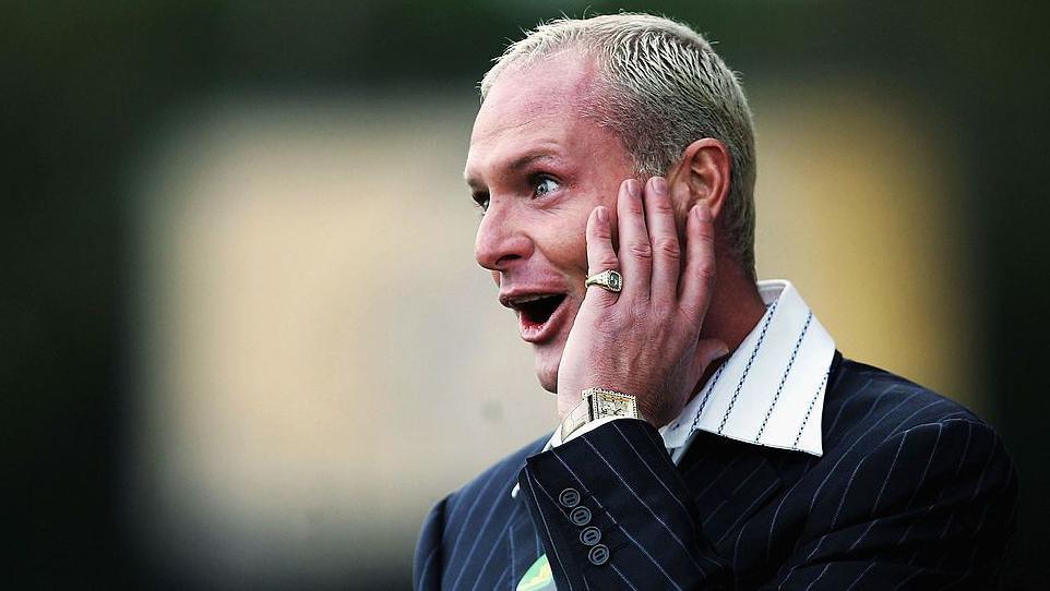 Paul Gascoigne reacting during a match as manager of Kettering Town. He had bleached hair and is wearing a dark pinstriped suit with a wide collared white shirt. He is holding his left hand to his face and gold watch is on his wrist.