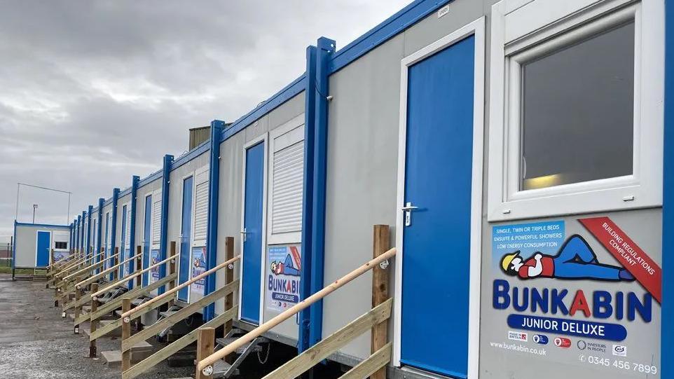 External view of the row of ten pods, grey portable cabins with wooden steps up to a blue front door