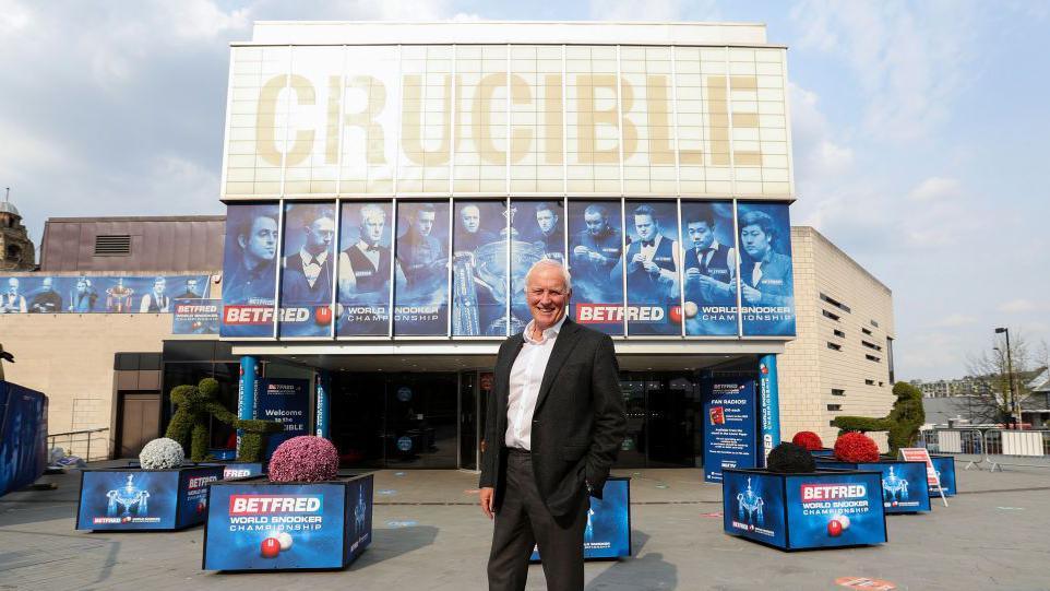 A man wearing a black suit and white shirt with short white hair stands outside the Crucible in Sheffield
