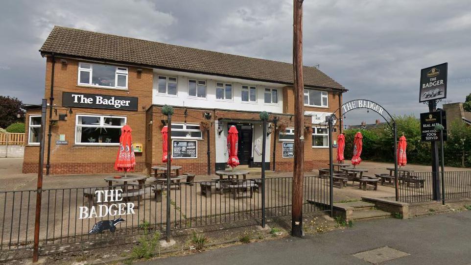 A 1960s-style pub with seating outside with red umbrellas. There are multiple signs which read THE BADGER.
