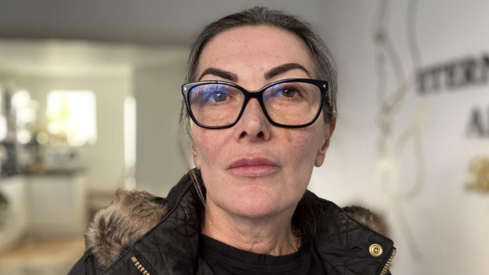 A woman in black framed glasses and a parka coat looking at the photographer. The photograph is a close up of her face but a room with a kitchenette area appears out of focus in the background. Her hair is tied back.