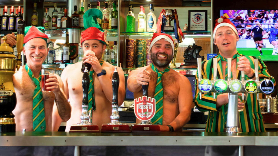 Four men in a Father Christmas hat, green and yellow-striped ties, are standing behind a bar table.