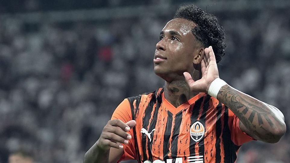 Kevin puts his hand to his ear in an orange and black striped Shakhtar shirt
