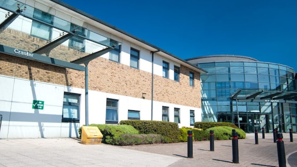 The outside of St George's Park hospital. There is a glass building to the right and a brick one to the left. Black bollards are on a paved area.