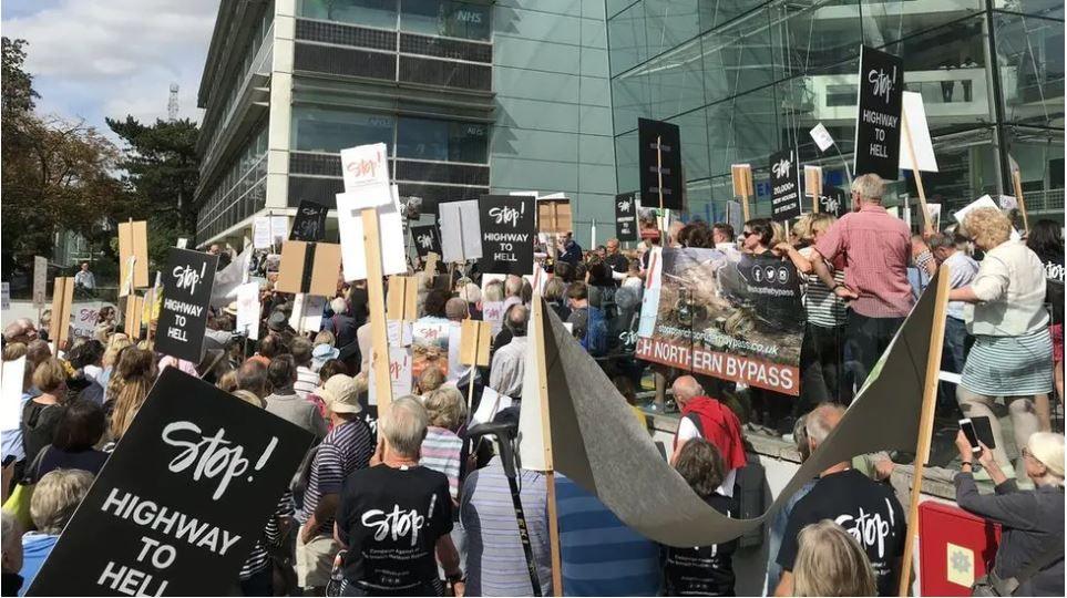 Many protestors outside the county council's headquarters, holding placards. Some say "Stop! Highway to hell."