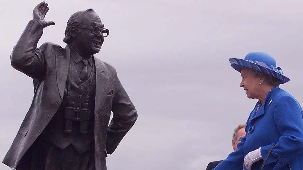 Queen Elizabeth II looks on at a statue of Eric Morecambe smiling while holding his hand aloft.